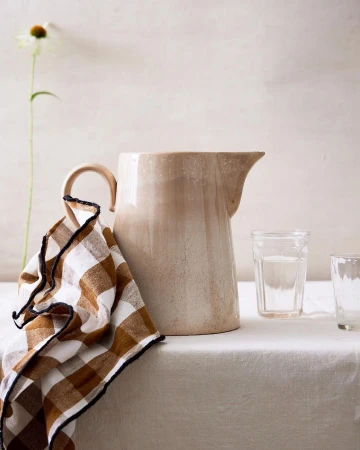 Still life photograph featuring a beige ceramic pitcher, two clear glasses, a checkered brown and white towel, and a white flower.