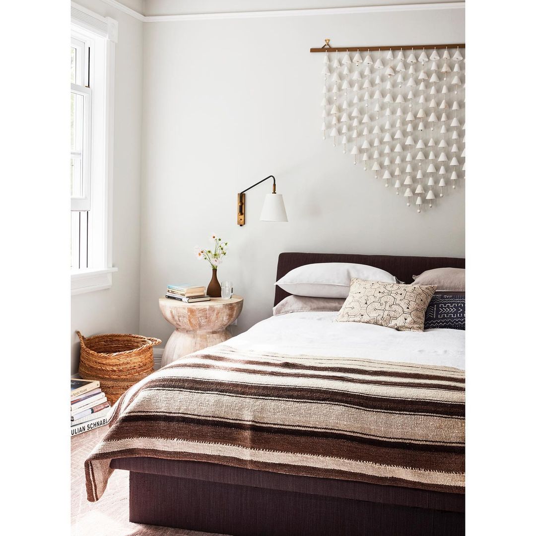 Bedroom interior featuring a bed with striped blanket, pillows, bedside table with books and flowers, and a woven wall decoration.
