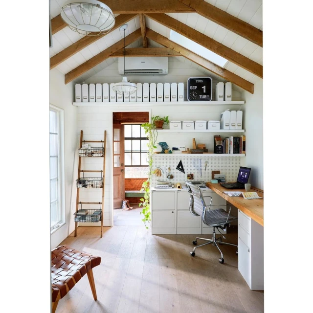 Home office interior with light wood floors, a woven leather chair, ladder shelf, white walls, desk, laptop, and a rustic wooden desk.