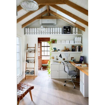 Home office interior with light wood floors, a woven leather chair, ladder shelf, white walls, desk, laptop, and a rustic wooden desk.