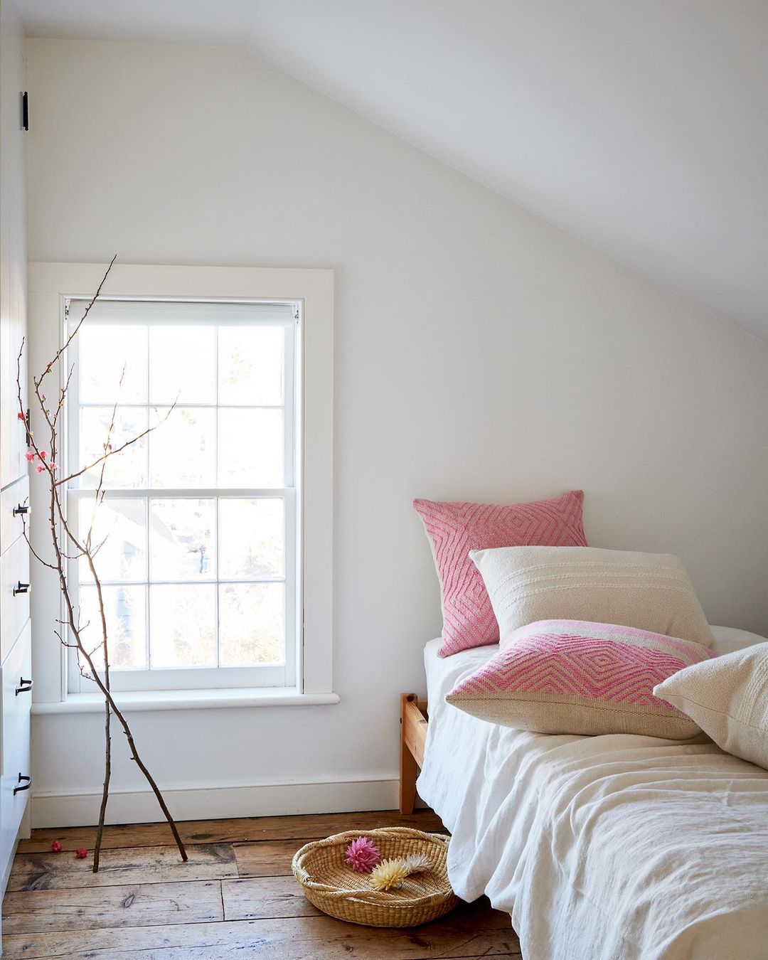 Bedroom interior with a wooden bed, white linens, pink pillows, bare branches in a vase, and a woven basket on a wooden floor.