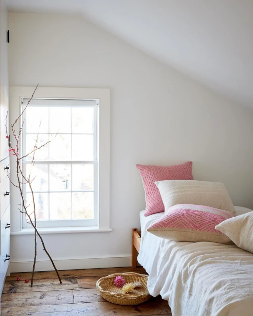 Bedroom interior with a wooden bed, white linens, pink pillows, bare branches in a vase, and a woven basket on a wooden floor.