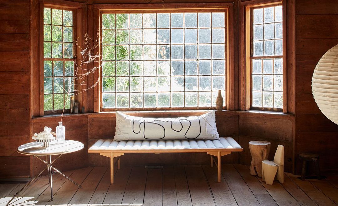 Indoor living space with light wood floors, a large grid window, and minimal furniture including a bench, table, vase, and lighting.