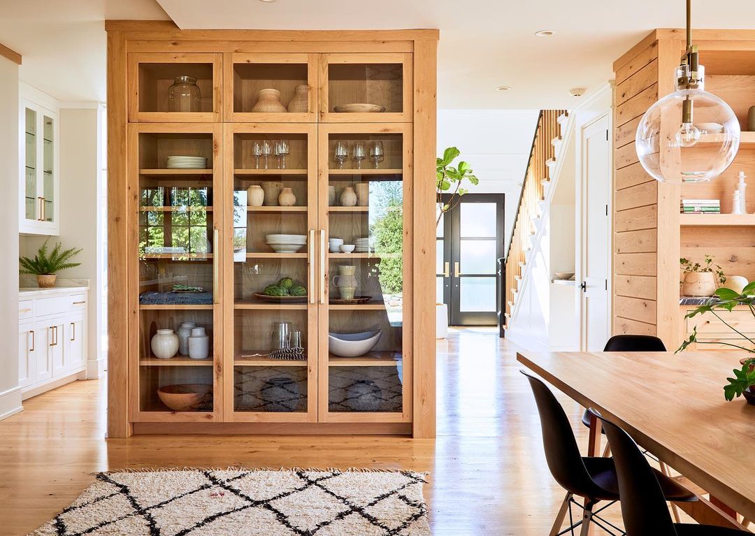 Bright and spacious home interior with a large wooden cabinet showcasing dishware, an area rug, and a wooden dining table.