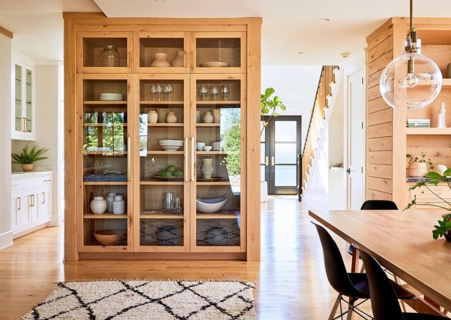 Bright and spacious home interior with a large wooden cabinet showcasing dishware, an area rug, and a wooden dining table.