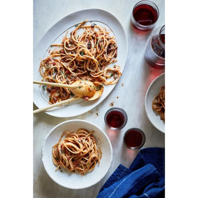 Overhead shot of two servings of spaghetti with tomato sauce in white bowls and a platter with wine glasses, featuring wooden serving spoons.