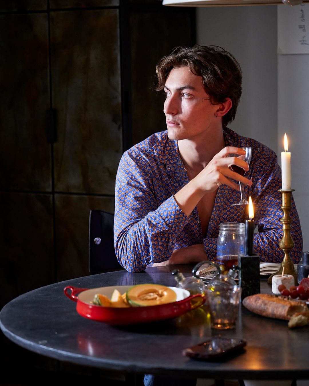 Dark hair man sits at table in patterned shirt. He holds wine glass and faces candles, fruit, and bread in an indoor setting.