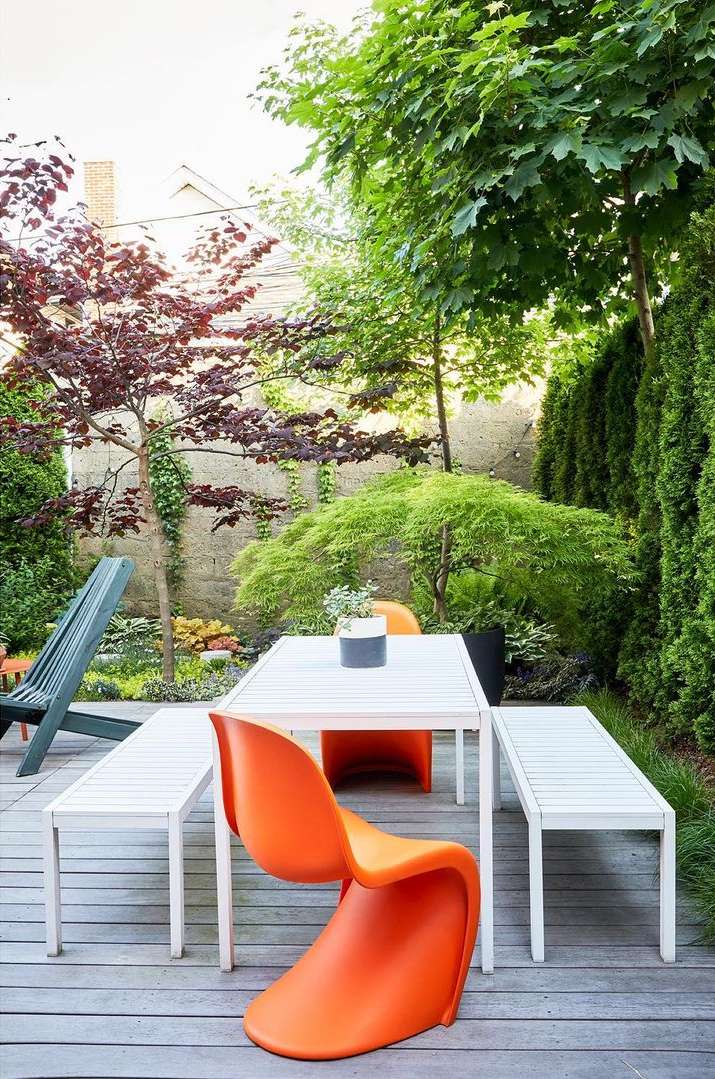 Outdoor patio with modern white table, benches, orange chair, and lush greenery. Perfect for dining or relaxation in a backyard setting.