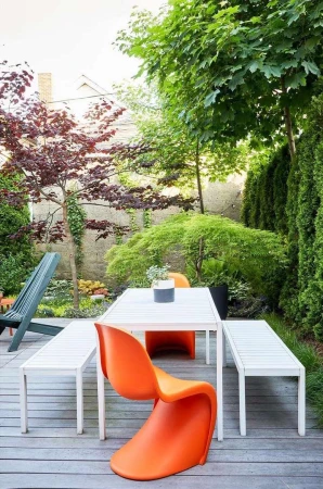 Outdoor patio with modern white table, benches, orange chair, and lush greenery. Perfect for dining or relaxation in a backyard setting.
