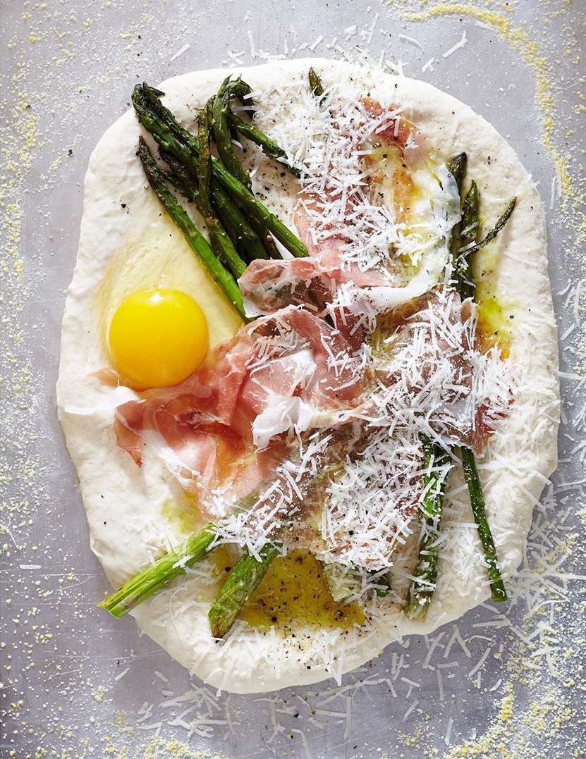 Overhead shot of an uncooked pizza topped with a raw egg yolk, asparagus, prosciutto, and parmesan cheese on a floured metal surface.