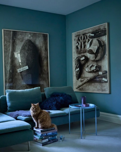 Interior scene with a ginger cat perched on a stack of books near a teal sectional sofa, complemented by modern art and decor.