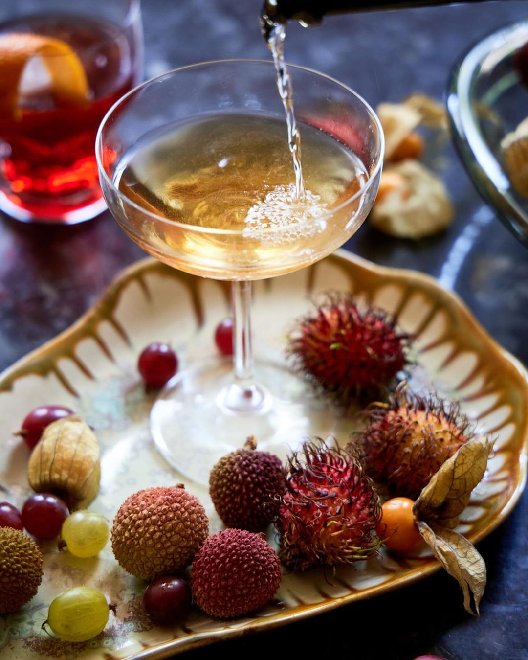 Champagne cocktail being poured into a coupe glass on a platter with rambutan, lychee, grapes, and golden berries in a studio.