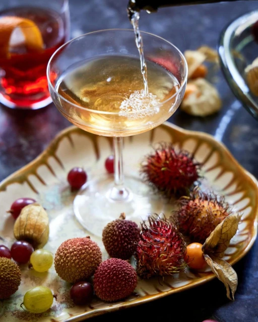 Champagne cocktail being poured into a coupe glass on a platter with rambutan, lychee, grapes, and golden berries in a studio.