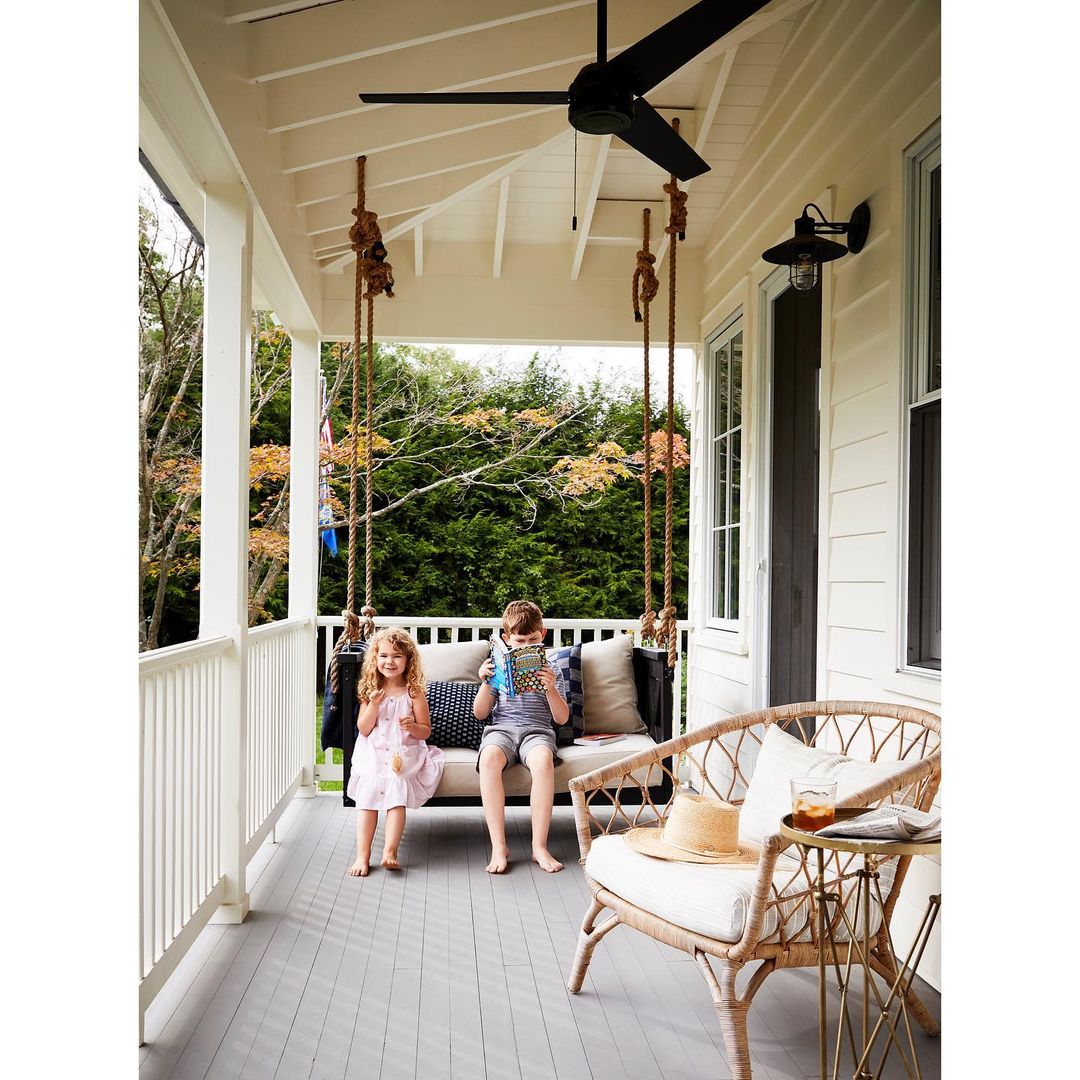 Two children, a boy and a girl, are sitting on a rope swing on a porch. The boy is reading a book. A rattan chair sits nearby.