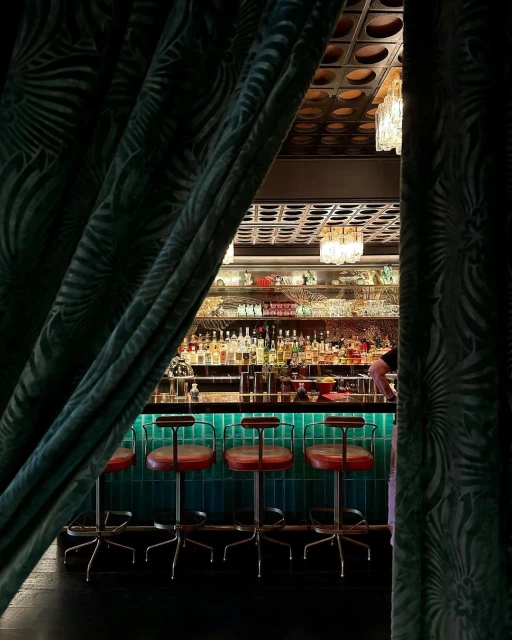 Interior view of a bar area with red bar stools lined along a turquoise counter, seen partially framed by dark green patterned curtains.