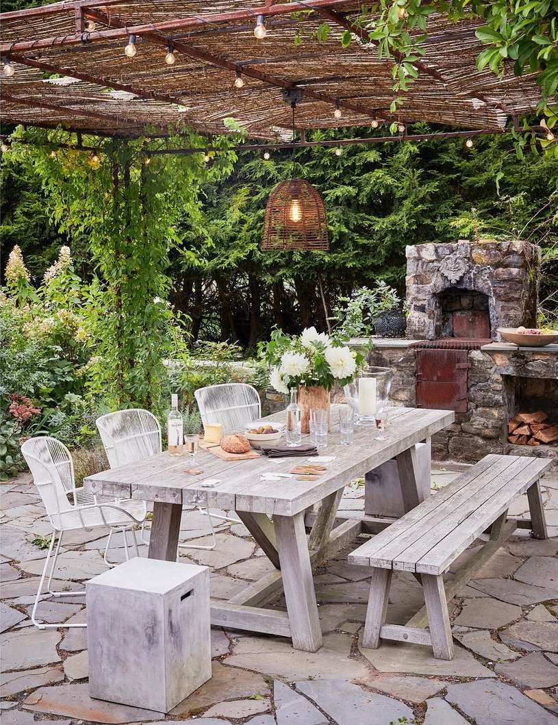 Outdoor dining area features a wooden table with chairs and bench, set under a woven pergola with string lights in lush garden.