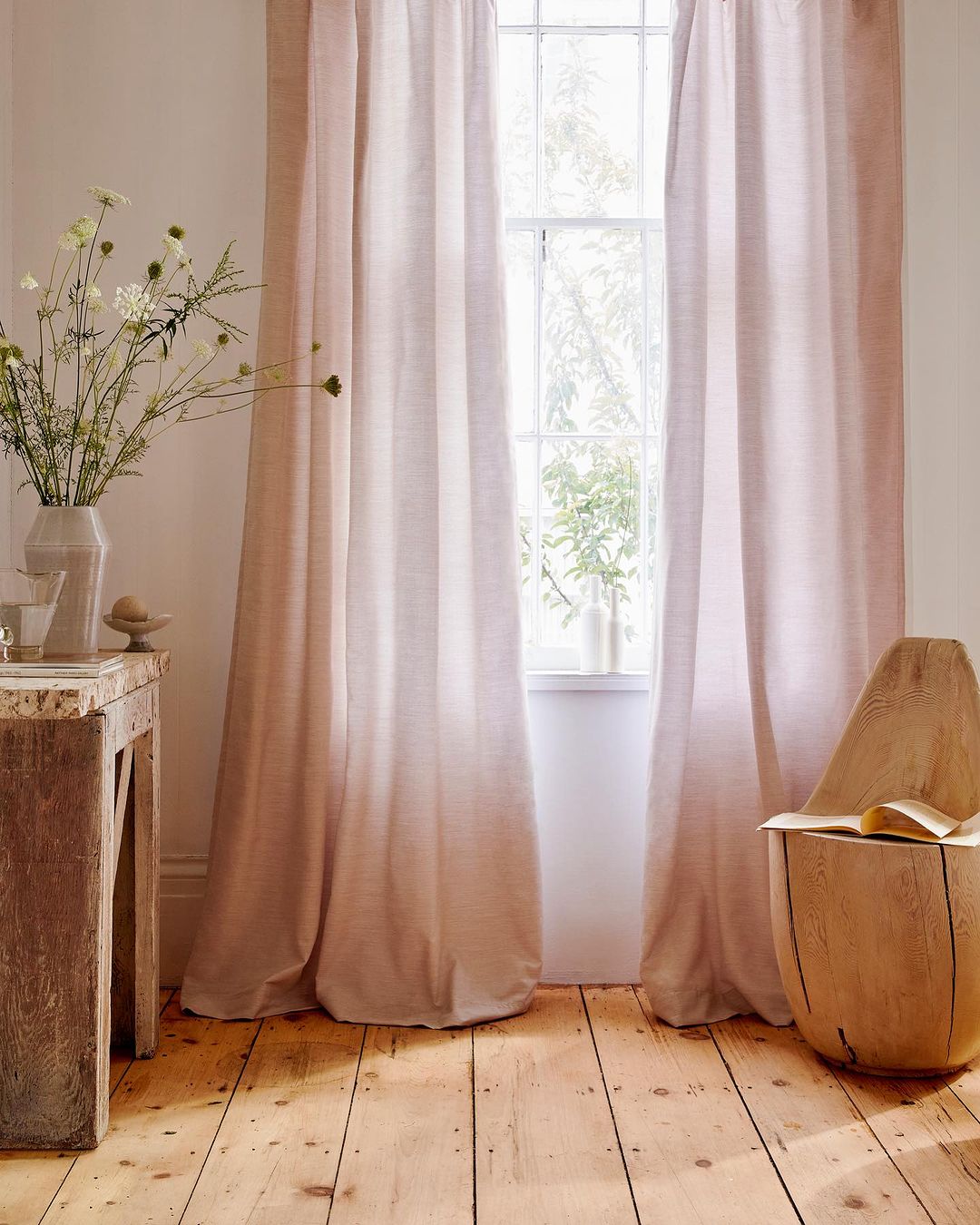 Interior view with pink linen curtains covering a window, light wood floors, a wooden side table, and a vase with white wildflowers.