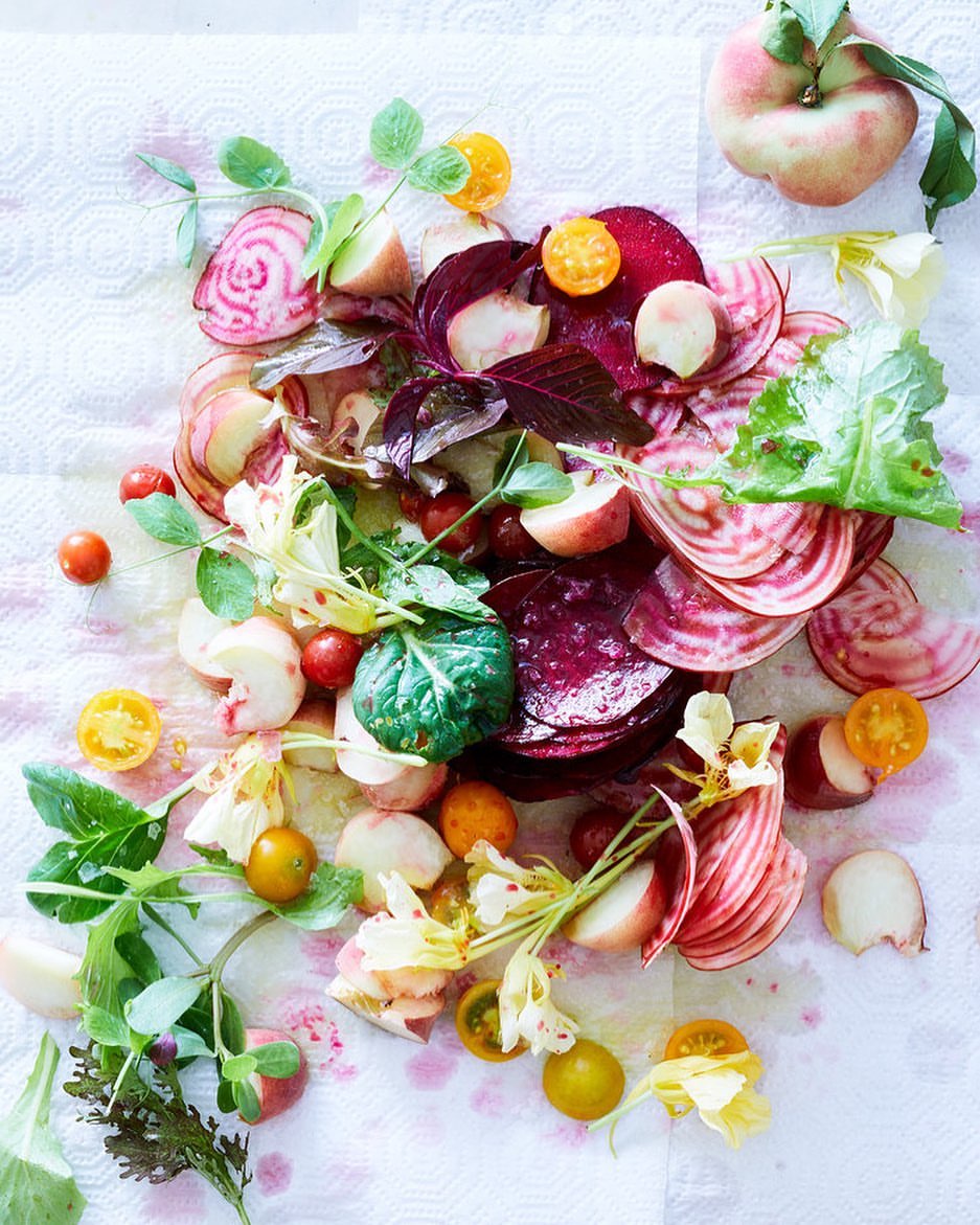 Overhead shot of sliced beets, peaches, cherry tomatoes, and greens on textured white surface, showcasing vibrant colors and textures.