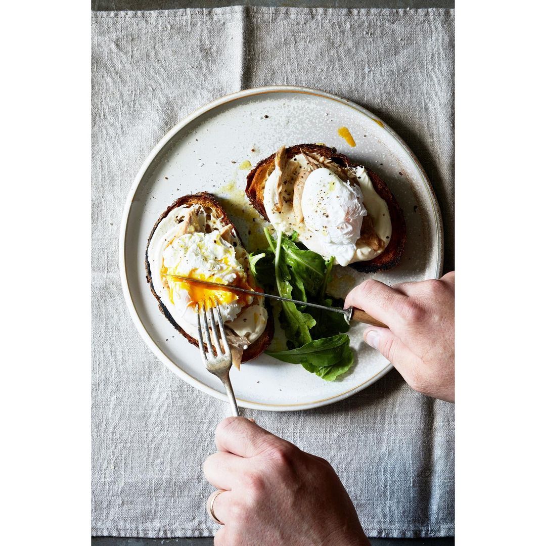 Person using knife and fork to cut open poached egg on toast with fresh greens. Breakfast food photography in natural light.