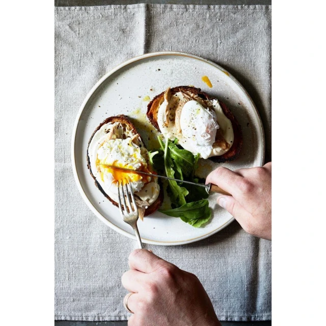 Person using knife and fork to cut open poached egg on toast with fresh greens. Breakfast food photography in natural light.