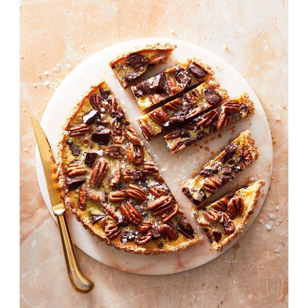 Overhead shot of a sliced pecan tart with chocolate chunks arranged on a round marble board with a gold knife. Food photography.