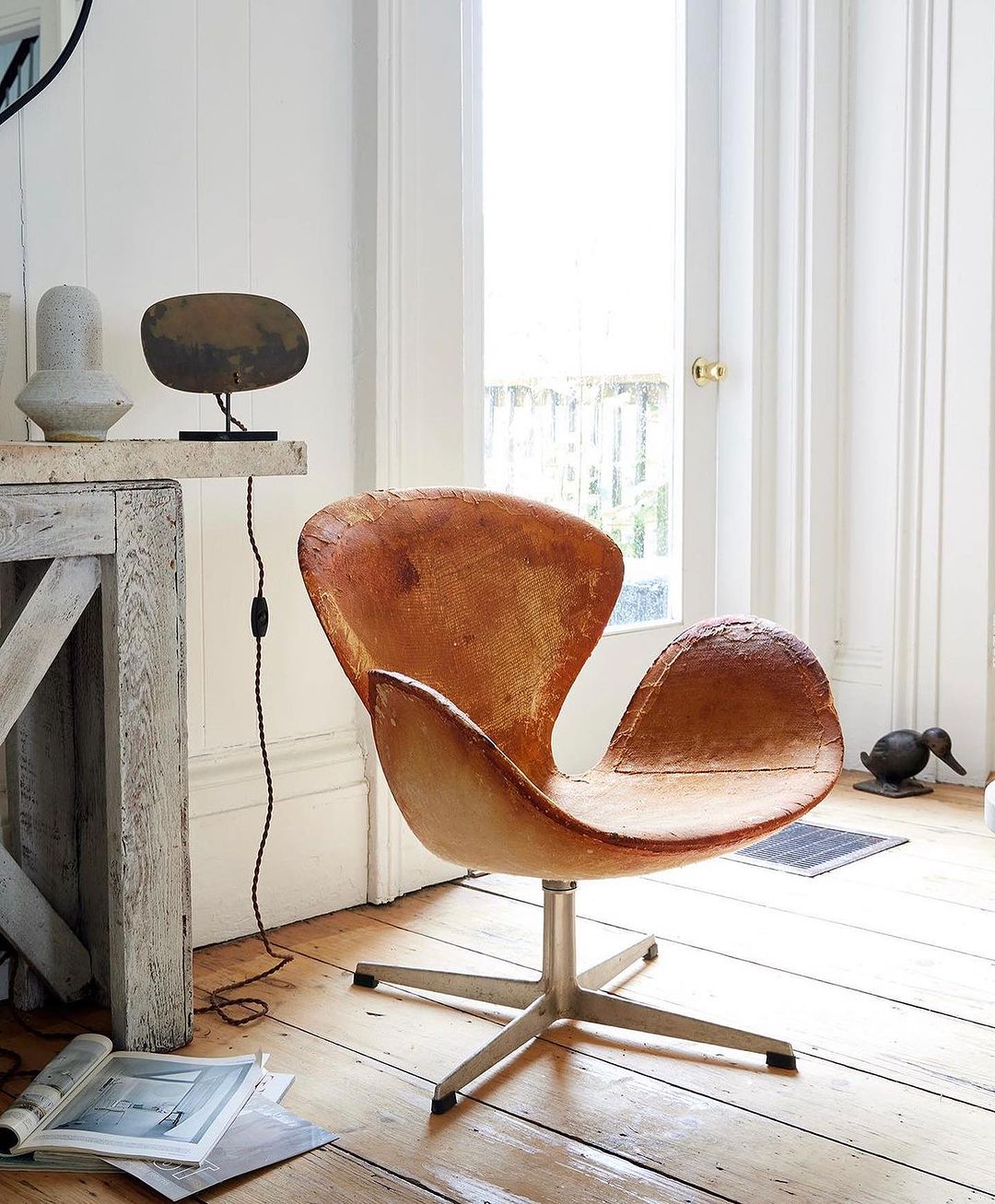 Arne Jacobsen Swan chair upholstered in tan leather with a metal swivel base, positioned on a wood floor in front of a window.