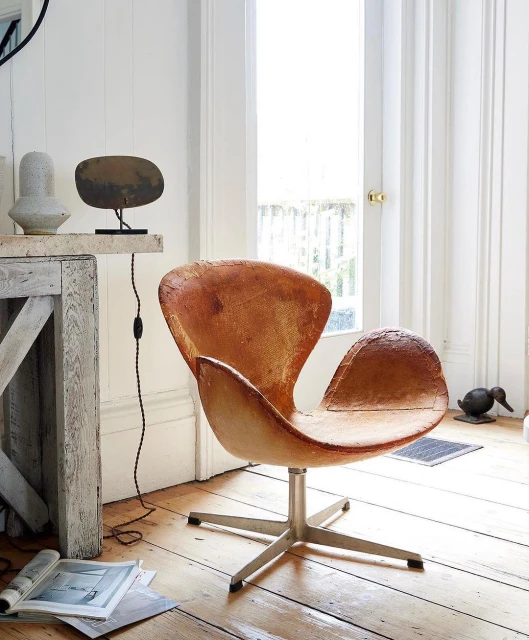 Arne Jacobsen Swan chair upholstered in tan leather with a metal swivel base, positioned on a wood floor in front of a window.