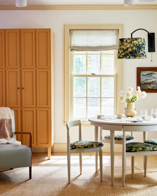 Neutral-toned dining room featuring a round table with chairs, a cabinet, and natural light from a window, creating a calm and inviting atmosphere.