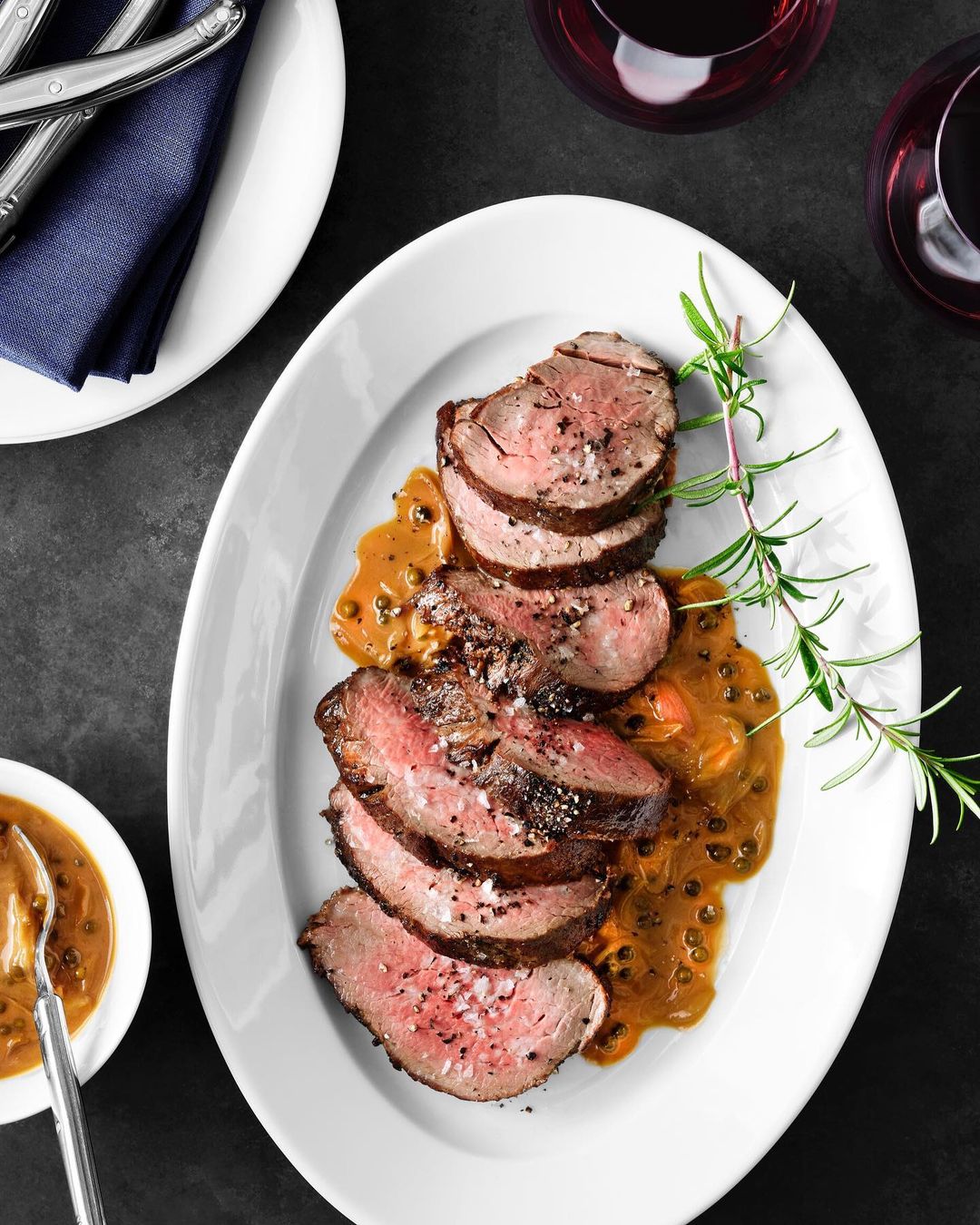 Overhead shot of sliced steak with peppercorn sauce on a white plate, accompanied by a bowl of sauce, wine glasses, and silverware.