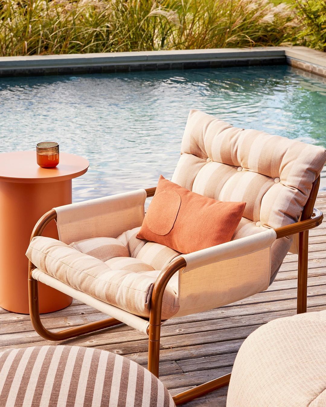 Outdoor lounge area featuring a cushion-covered chair, round side table with a drink, striped ottoman, and swimming pool in the background.