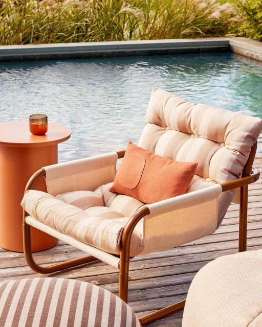Outdoor lounge area featuring a cushion-covered chair, round side table with a drink, striped ottoman, and swimming pool in the background.