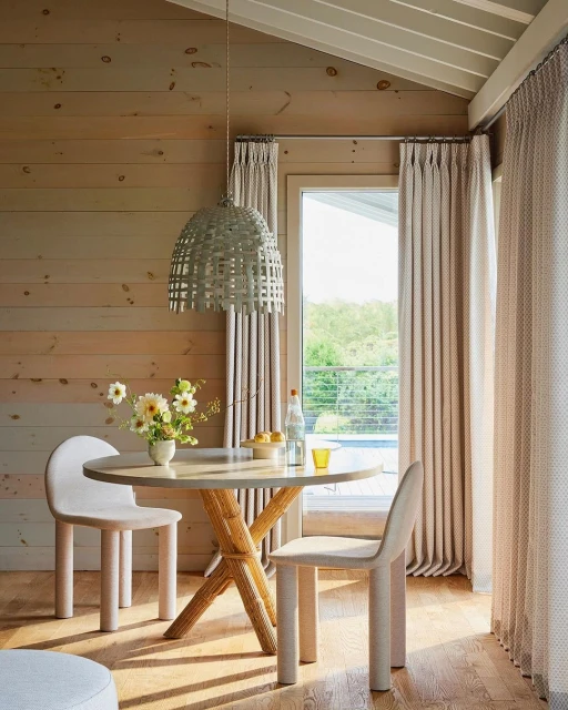 Coastal dining room with round table, straw pendant light, and two chairs against a backdrop of wood paneling and patterned curtains.