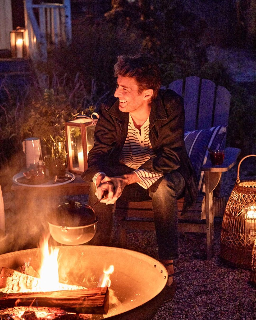 Man sits by a fire pit outdoors at night, with glowing lanterns nearby. He wears jeans and a striped shirt, smiling and relaxed in the warm light.