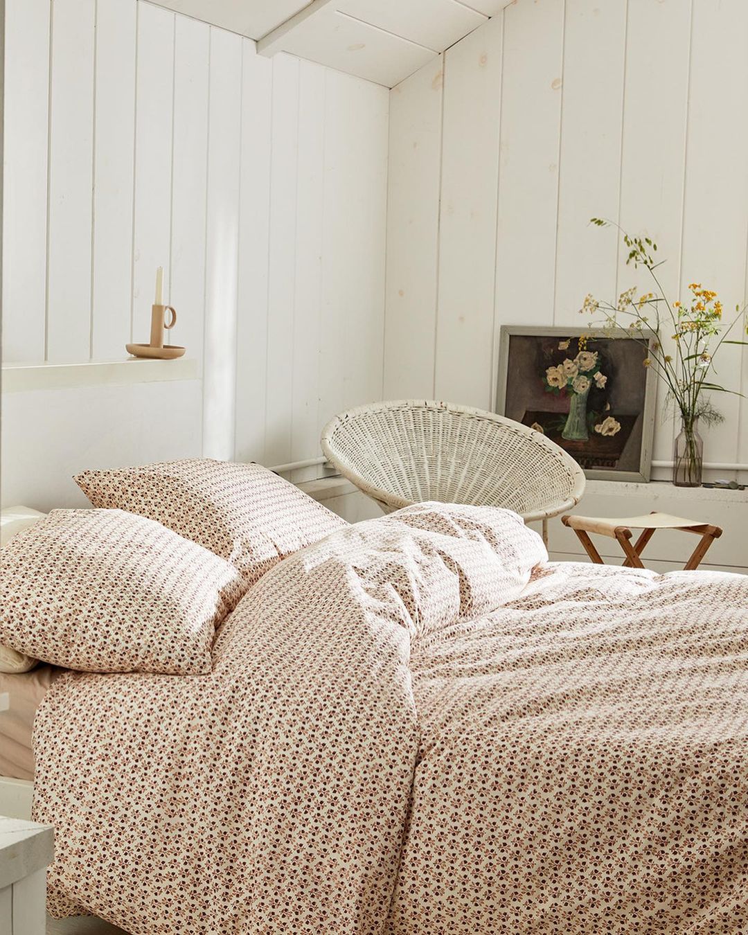 Bedroom interior with patterned bedding, wicker chair, painting, and wildflowers in a bottle, creating a cozy and inviting atmosphere.