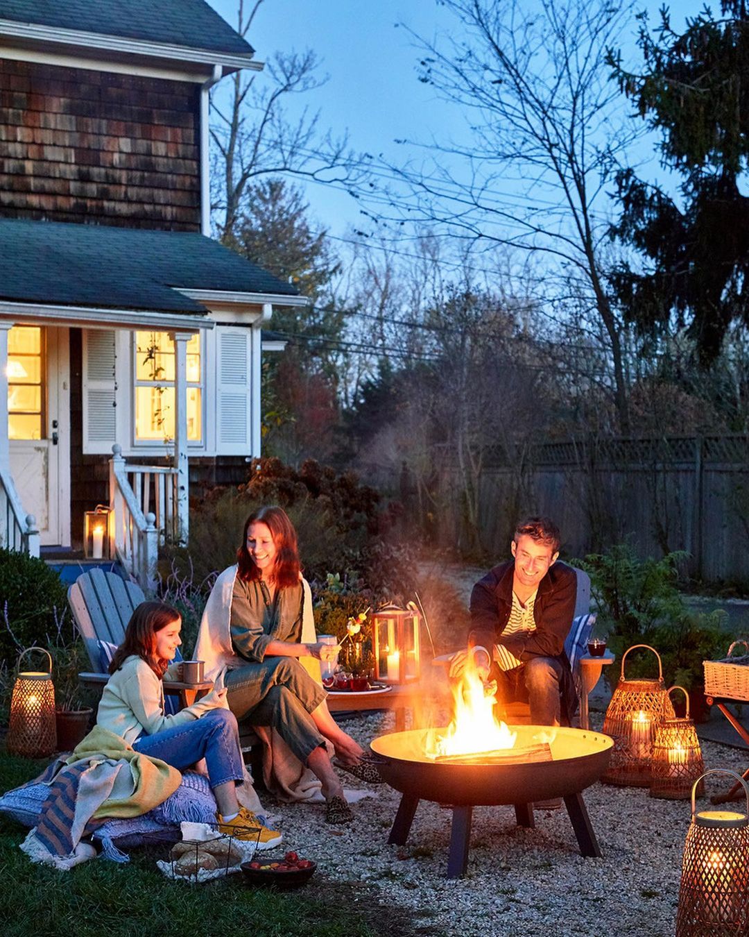 Three people sitting around a fire pit in a backyard at dusk, with a house and trees in the background, and lanterns scattered around.