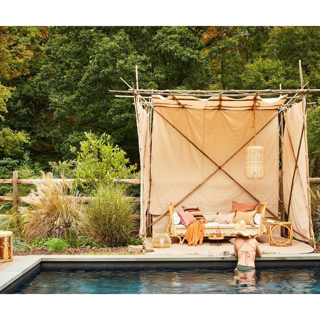 Back view of adult female wading in a pool towards a poolside lounge area shaded by an outdoor tent structure in a backyard.