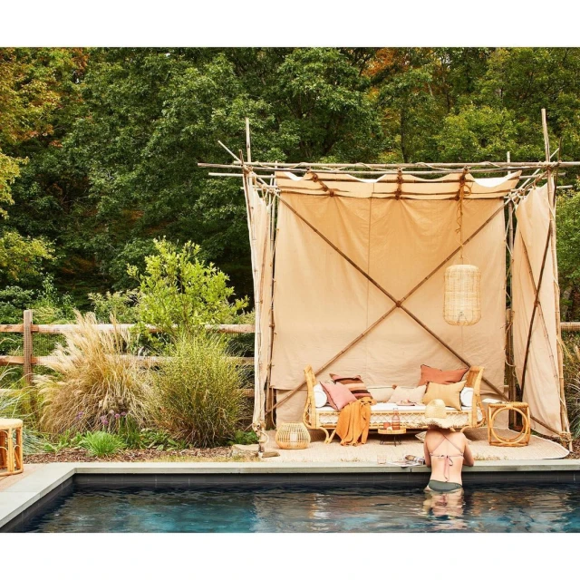 Back view of adult female wading in a pool towards a poolside lounge area shaded by an outdoor tent structure in a backyard.