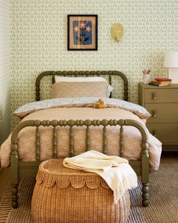 Child's bedroom features a green metal bed frame with floral bedding, woven storage ottoman, wall art, and a sage green dresser.