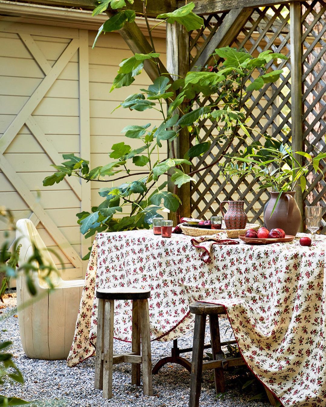 Outdoor garden table set with floral tablecloth, glasses, plums, and wooden stools on gravel. Trellis and leafy tree in background.
