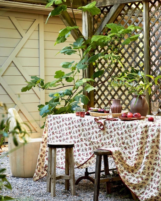 Outdoor garden table set with floral tablecloth, glasses, plums, and wooden stools on gravel. Trellis and leafy tree in background.