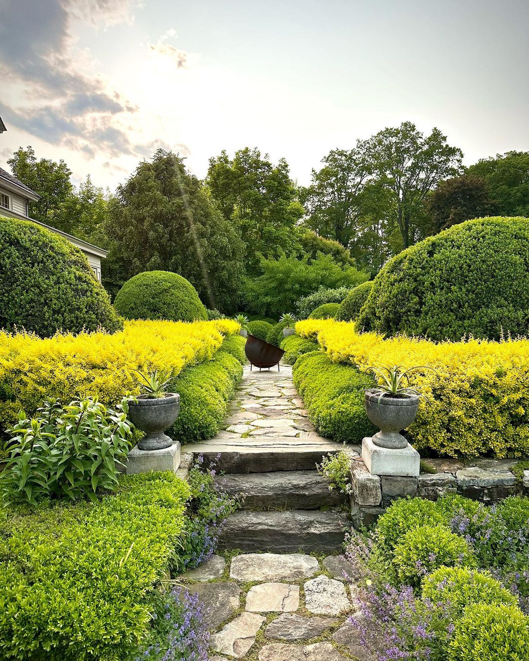 Landscaped garden with stone walkway, urn planters, and manicured bushes in yellow and green under a cloudy sky, offering serene beauty.