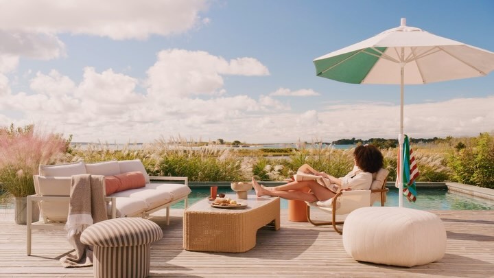 Woman relaxes outdoors in lounge chair by pool with furniture, umbrella, and coastal view under blue sky with clouds in daylight.