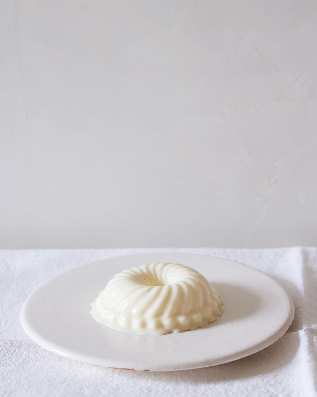 A close-up shot of a round, white dessert with a ridged design, displayed on a white plate and a white tablecloth against a white background.