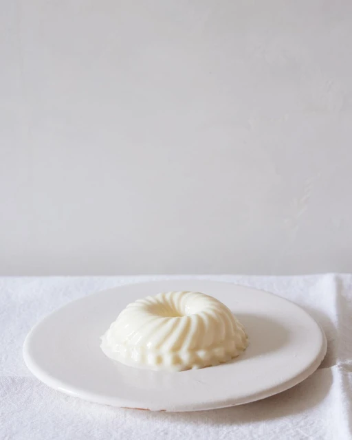 A close-up shot of a round, white dessert with a ridged design, displayed on a white plate and a white tablecloth against a white background.