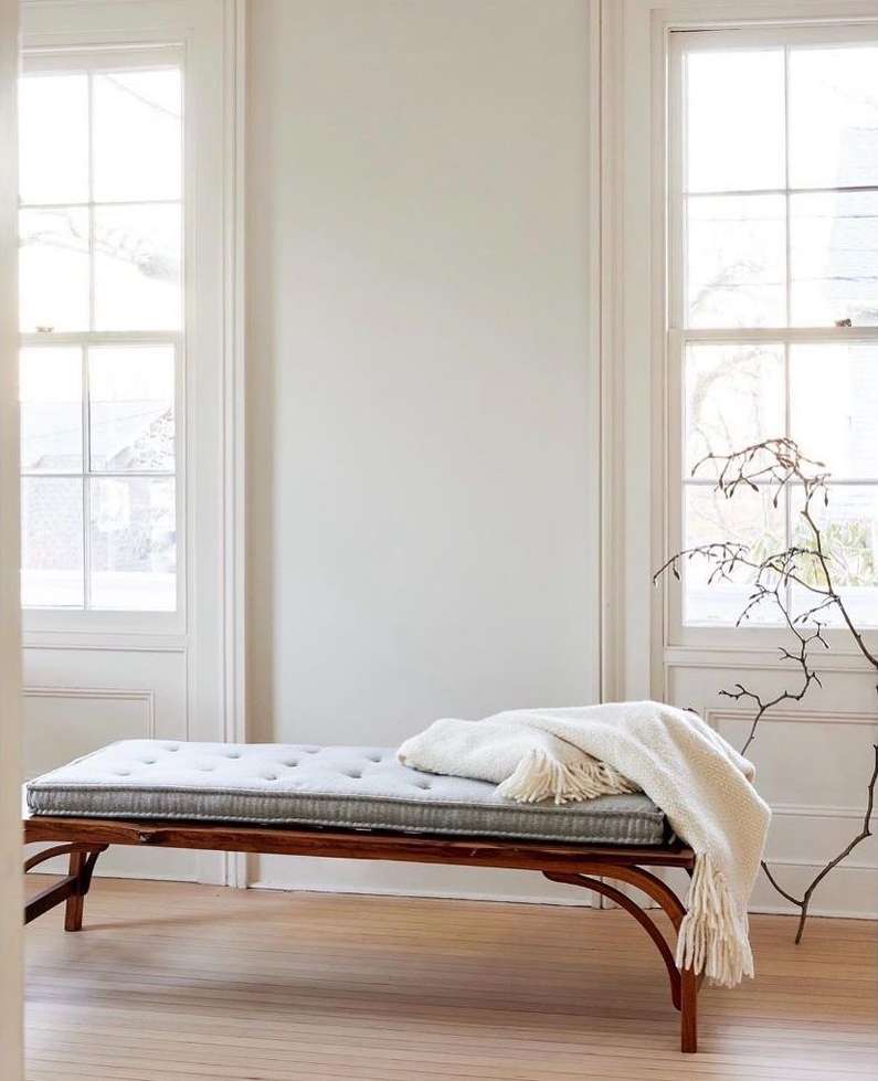 Interior of room featuring a daybed with a gray cushion and a white blanket, set against a backdrop of white walls and windows.