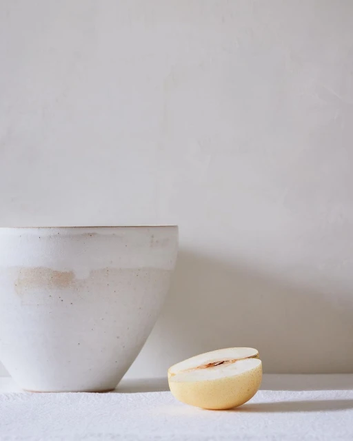 Close-up of a white ceramic bowl beside a halved Asian pear on a bright white surface against a light neutral background. Food photography.