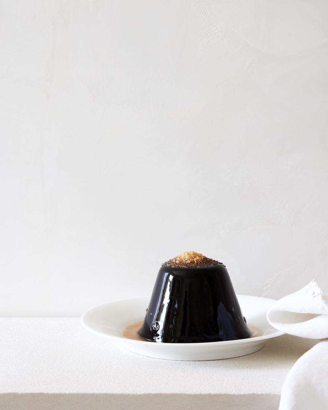 Elegant black jelly dessert topped with brown sugar, served on a white plate with a cloth napkin, against a muted white background.