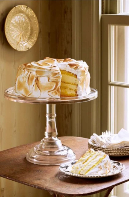 Lemon meringue cake on a glass stand next to a slice on a plate, and napkins on a table in a room with tan walls and natural light.