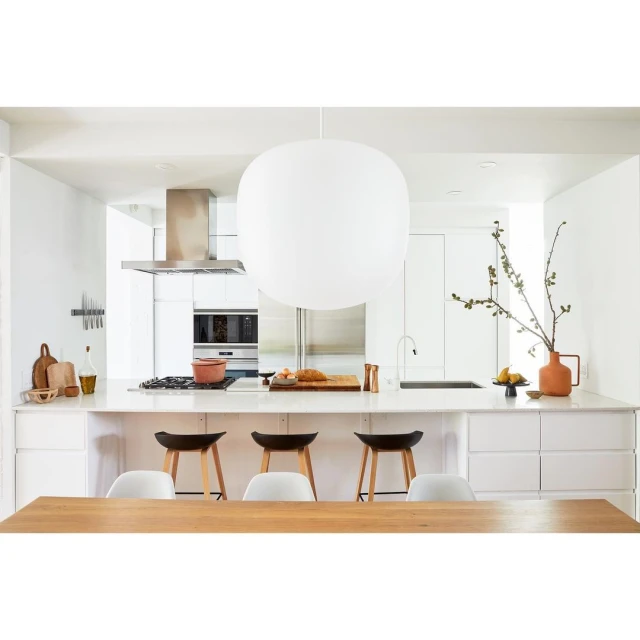 Bright, modern kitchen interior with island, white cabinets, overhead lighting, stools, hardwood table, and vase with branch.