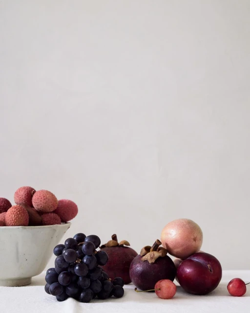 Still life of fruits including grapes, lychees, mangosteens, plums, and a guava in a white bowl, set against a neutral backdrop.